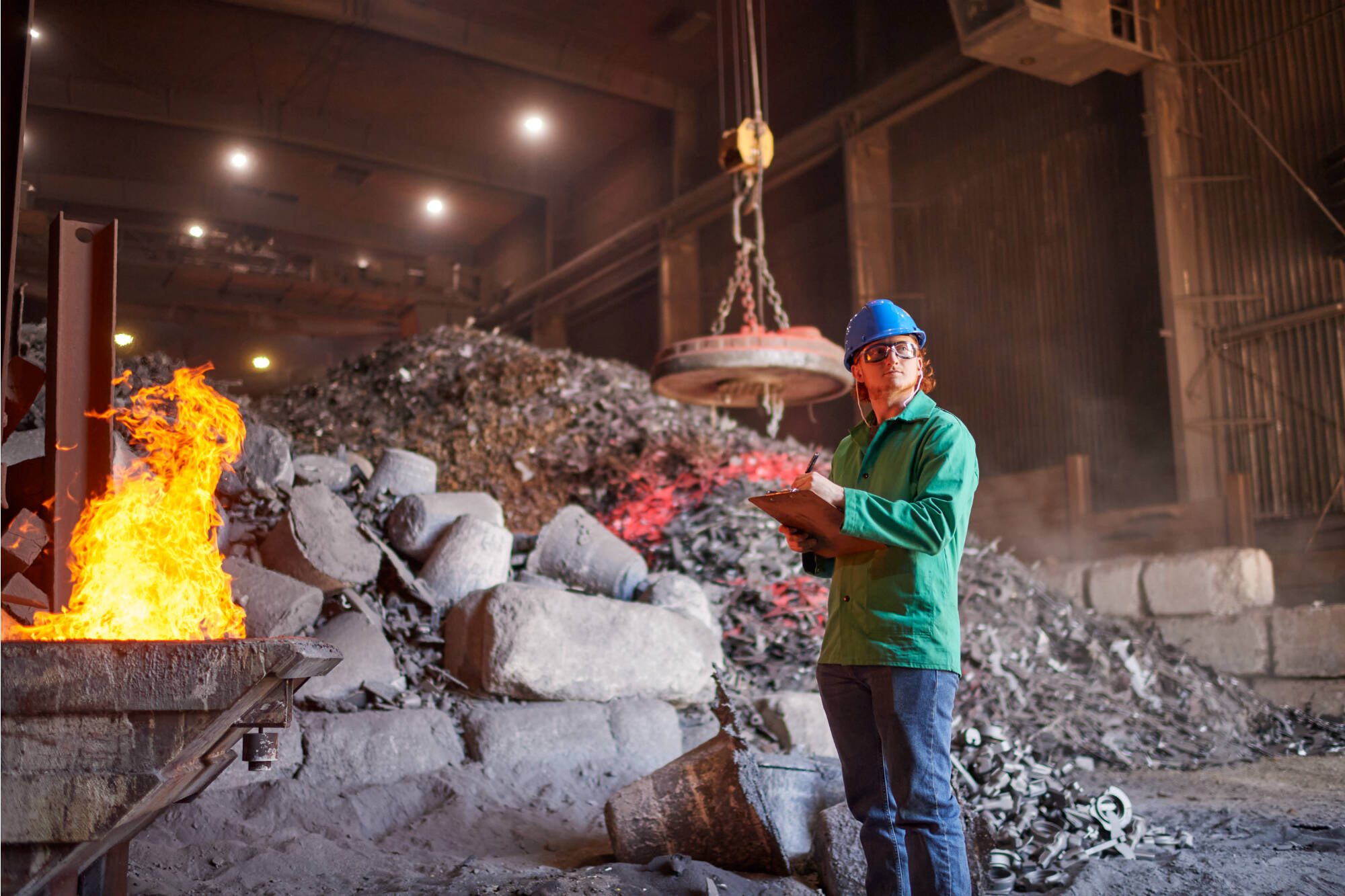 A worker in a blue hard hat and green shirt stands in a scrapyard, holding a clipboard and pen, near a burning pile of metal.